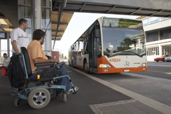 Ein Rollstuhlfahrer und eine stehende Person warten in der Station auf den herannahenden Bus.