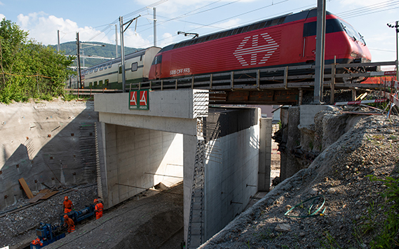 A red locomotive at the head of a double-decker train passes over a new bridge above a construction site.