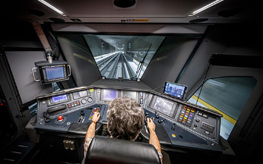 Vue de l’intérieur d’une cabine de conduite d’une locomotive moderne qui traverse un tunnel. Plusieurs écrans, des voyants lumineux et des boutons de plusieurs couleurs délimitent le plan de travail du conducteur de locomotive. Le véhicule moteur fait partie d’une rame Flirt du Léman Express.