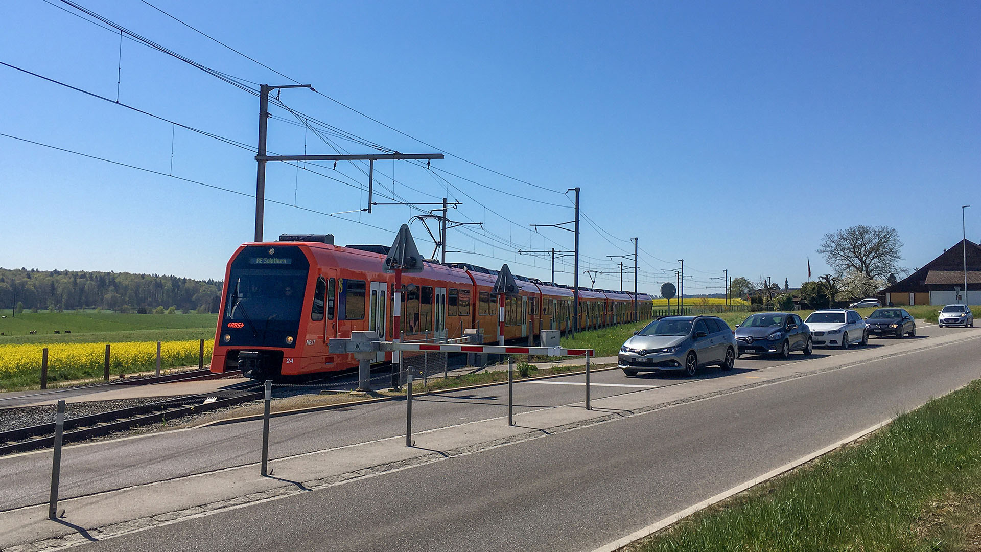 Un treno arancione della RBS e una colonna di auto ferme davanti a una barriera.